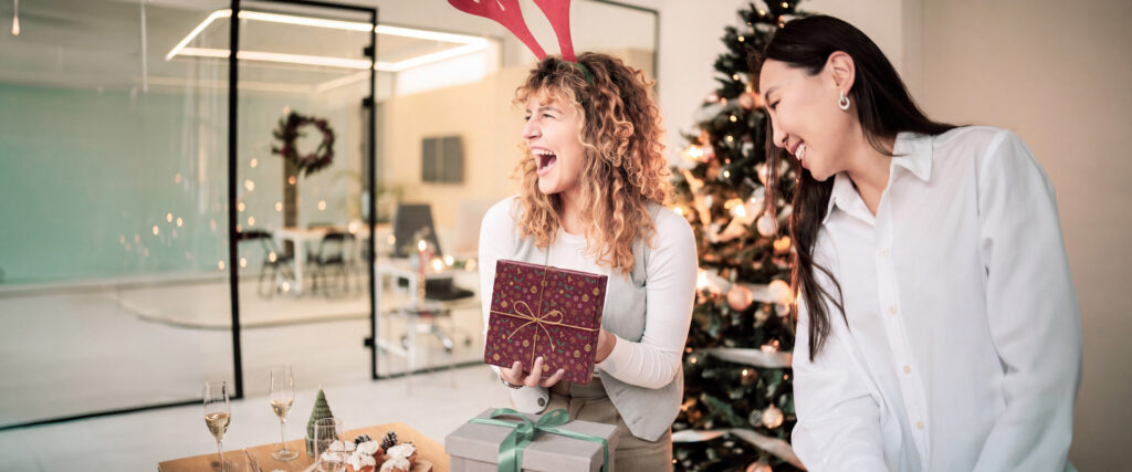 Zwei Frauen im Office, die Weihnachtsgeschenke auf einen Office Tisch legen. Im Hintergrund ist das Office weihnachtlich geschmückt.