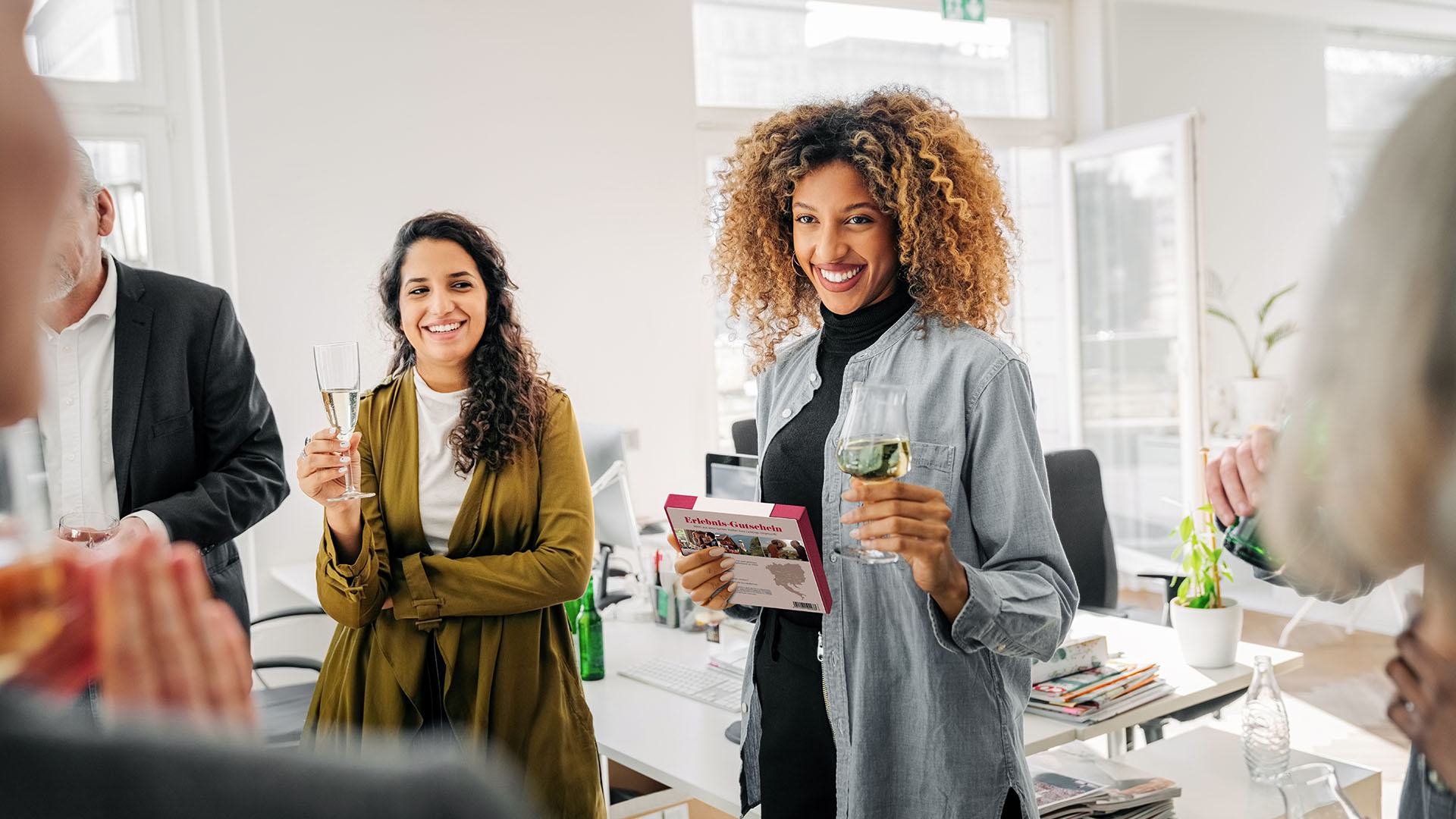 Das Bild zeigt eine Gruppe von Menschen, die an einem scheinbar festlichen Anlass in einem modernen Büro oder einem ähnlichen Arbeitsumfeld teilnehmen. Zwei Frauen stehen im Vordergrund, beide halten ein Glas in der Hand und lächeln fröhlich. Die Frau rechts hat lockiges, blondes Haar und trägt ein graues Hemd über einem schwarzen Rollkragenpullover. In ihrer linken Hand hält sie ein Geschenk oder eine Karte. Die Frau links hat langes, dunkles, lockiges Haar und trägt eine grüne Jacke über einem weißen Oberteil. Im Hintergrund sind weitere Personen zu sehen, die ebenfalls an der Feier teilnehmen. Die Umgebung wirkt hell und freundlich, mit viel Tageslicht, Pflanzen und Büromöbeln im Hintergrund. Es scheint, dass die Menschen miteinander anstoßen und eine angenehme Zeit miteinander verbringen.